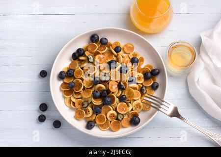 Teller Mini Pancakes Müsli mit Honig, Beeren und frischem Orangensaft auf hellblauem Hintergrund. Trendige Küche. Hausgemachtes Frühstück. Draufsicht. Platz Stockfoto