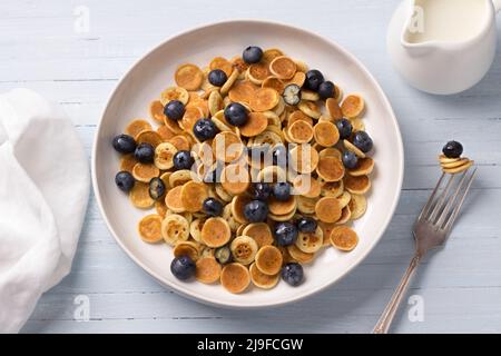 Teller Mini Pancakes Müsli mit Creme, Honig und Beeren auf einem hellblauen Hintergrund. Trendige Speisen. Hausgemachtes Frühstück. Draufsicht Stockfoto