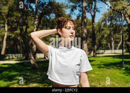 Rotschopf schöne Frau trägt weißes T-Shirt, hält Haare und schaut weg, während sie auf dem grünen Stadtpark im Freien steht. Stockfoto