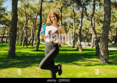 Rotschopf wunderschöne Frau, die Sport-BH und schwarze Leggings trägt, eine Flasche Power Drink hält und vor der Kamera posiert, während sie draußen auf dem grünen Stadtpark steht Stockfoto