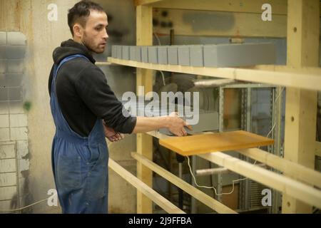 Der Arbeiter stellt die Schachtel auf das Regal. Mann faltet bemalte Bretter. Guy arbeitet in der Holzwerkstatt. Stockfoto