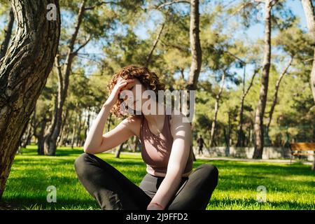 Attraktive Rothaarige Frau, die im Park auf dem Gras sitzt und starke Kopfschmerzen hat. Sie berührte ihre Schläfe und spürte Stress oder Schmerzen. Stress oder Kopfschmerzen concep Stockfoto