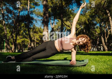 Junge Rotschopf sportliche Frau versucht Vasisthasana Seitenplanke, Arm Bein Unterstützung balancierende Pose auf grünen Stadtpark, im Freien. Gesunder Lebensstil, Outdoor sp Stockfoto