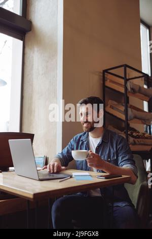 Fröhlicher kaukasischer junger attraktiver Mann mit einer Tasse Kaffee, der im Café am Laptop sitzt. Videoanruf. Stockfoto