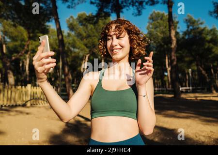 Junge Rotschopf Frau trägt Sport-BH auf Stadtpark, im Freien Telefon halten Blick auf Bildschirm winkende Hand Video Anruf Distanz Freund online in mobi Stockfoto