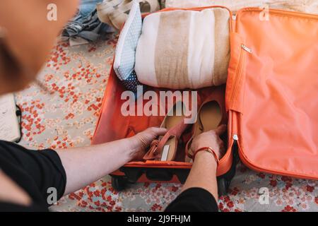 Nahaufnahme eines Koffers, der für einen Sommerurlaub verpackt wird. Hände einer Frau, die Schuhe in ihr Gepäck legt. Reise- und Urlaubskonzept Stockfoto