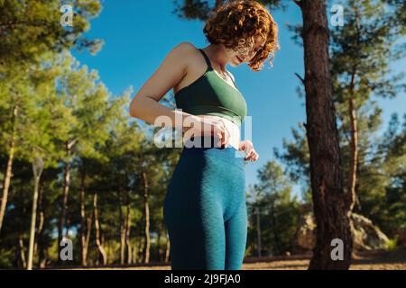 Nette Rothaarige Frau trägt grünen Sport-BH und blaue Yoga-Hosen stehen auf Stadtpark, im Freien hält sie die Hände an der Taille. Flacher Magen, gesund li Stockfoto