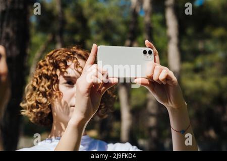 Junge nette Rothaarige Frau in weißem T-Shirt auf grünen Stadtpark stehend, im Freien horizontale Foto mit ihrem Smartphone. Stockfoto