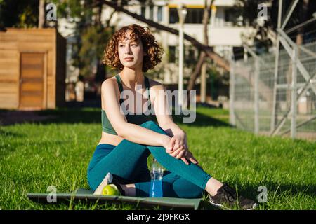 Junge Rothaarige Frau mit grünem Sport-BH und blauer Yogahose sitzt auf einer Yogamatte im Stadtpark und blickt im Freien zweifelhaft weg. Yoga und Stockfoto