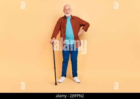 Foto von ziemlich lustig Kerl Rentner gekleidet braun Hemd Brille halten Gehstock isoliert beige Farbe Hintergrund Stockfoto