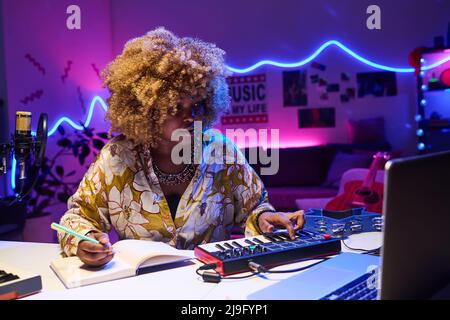 Horizontales mittleres Porträt einer jungen afroamerikanischen Frau, die zu Hause am Schreibtisch sitzt und mit Synthesizer und Laptop Songs komponiert Stockfoto