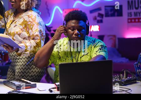 Moderner junger afroamerikanischer Mann mit Kopfhörern, der mit einer Sängerin im Aufnahmestudio arbeitet und die Toneinstellungen auf dem Laptop anpasst Stockfoto