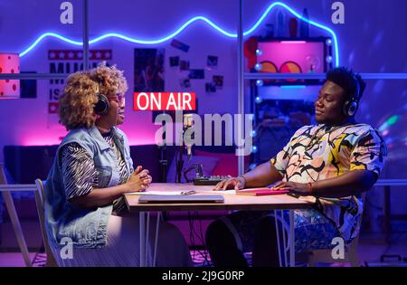Junge afroamerikanische Männer und Frauen sitzen vor dem Tisch und nehmen Podcast auf oder übertragen sie im Heimstudio Stockfoto