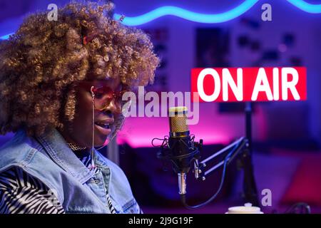 Mittlere Nahaufnahme einer jungen, eleganten afroamerikanischen Frau, die vor dem Mikrofon sitzt und Podcasts oder Sendungen aufnimmt Stockfoto