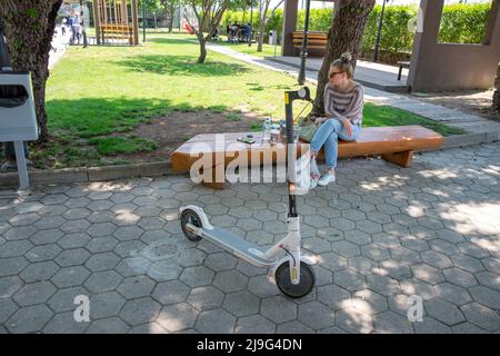 Frau sitzt auf einer Holzbank und genießt ihren Tag im Park mit ihrem Elektroroller im Vordergrund Stockfoto