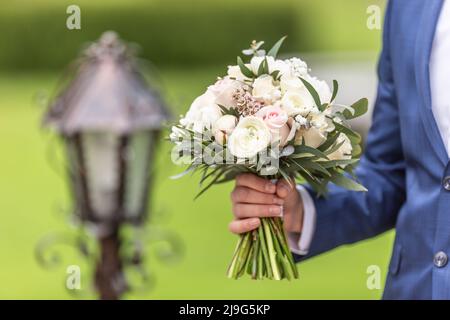 Der Bräutigam hält an einem Hochzeitstag einen Hochzeitsstrauß mit weißen Blumen im Freien. Stockfoto
