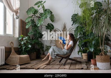 Junge, entspannte, verträumte Frau, die einen Papierventilator verwendet, während sie sich im schönen, erfrischenden Hausgarten entspannt Stockfoto