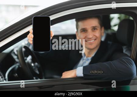 Glücklicher junger Autoverkäufer, der sein Smartphone mit leerem Bildschirm zeigt, während er im neuen Auto beim Autohändler sitzt, Modell Stockfoto