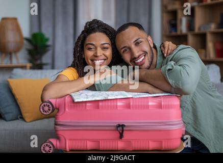 Positives afroamerikanisches Paar, das sich auf den Koffer lehnte und vor der Kamera lächelte, bereit für einen Urlaub im Ausland, freier Platz Stockfoto