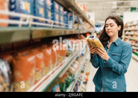 Einkaufen im Lebensmittelgeschäft. Eine junge Frau in einem Jeanshemd liest mit einem Lächeln die Informationen auf einem Päckchen Pasta. Im Vordergrund verschwommene Regale Stockfoto