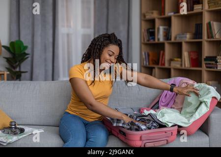 Konzept zur Reisevorbereitung. Glückliche schwarze Frau, die Koffer packte und sich für den Urlaub vorbereite, auf dem Sofa zu Hause sitzend Stockfoto