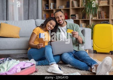 Glückliche afroamerikanische Ehepartner, die mit einem Laptop-Computer unterwegs waren und Fahrkarten kauften, sitzen mit ausgepackten Koffern auf dem Boden Stockfoto