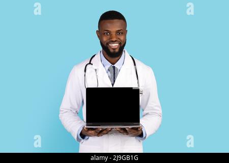 Lächelnder Schwarzer Doktor In Weißem Mantel Hält Laptop Mit Leerem Bildschirm Stockfoto