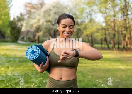 Fröhliche schwarze Frau, die die Smartwatch anschaut, die Fitnessaktivitäten verfolgt, eine Sportmatte hält, bereit für das Training im Freien Stockfoto