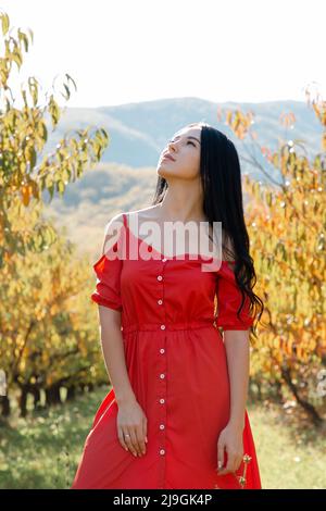 Spaziergang im Garten. Eine brünette Frau in einem roten Kleid posiert vor dem Hintergrund von Bäumen und blickt in den Himmel. Vertikale Ausrichtung. Stockfoto