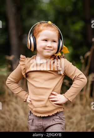 Ein kleines fröhliches Mädchen mit roten Haaren und Kopfhörern, das an einem Herbsttag im Park Musik hört Stockfoto