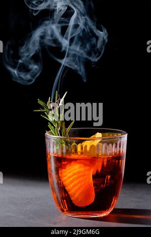 Negroni-Cocktail mit rauchenden Rosmarin- und Orangengarnitur auf schwarzem Hintergrund, minimalistischer Stil Stockfoto