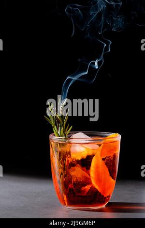 Negroni-Cocktail mit rauchenden Rosmarin- und Orangengarnitur auf schwarzem Hintergrund, minimalistischer Stil Stockfoto