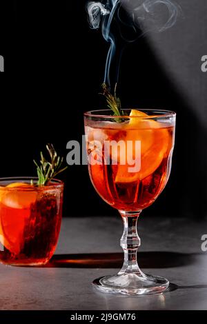 Negroni-Cocktail mit rauchenden Rosmarin- und Orangengarnitur auf schwarzem Hintergrund, minimalistischer Stil Stockfoto