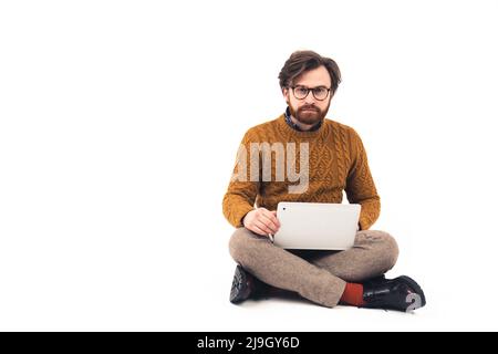 Der faszinierte kaukasische männliche Social-Media-Nutzer sitzt im Studio auf dem Boden mit einem Laptop auf den Knien auf weißem Hintergrund. Hochwertige Fotos Stockfoto
