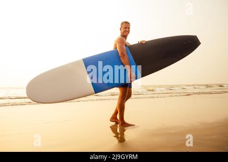 Surfen Freiberuflicher Mann Muskel-und Pressesporttraining am Strand bei Sonnenuntergang.männliche Fitness-Modell Surfer mit einem großen Surfbrett in der Nähe des indischen Ozeans Stockfoto