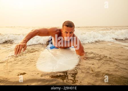 Surfen Freiberuflicher Mann Muskel-und Pressesporttraining am Strand bei Sonnenuntergang.männliche Fitness-Modell Surfer mit einem großen Surfbrett in der Nähe des indischen Ozeans Stockfoto