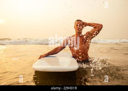 Surfen Freiberuflicher Mann Muskel-und Pressesporttraining am Strand bei Sonnenuntergang.männliche Fitness-Modell Surfer mit einem großen Surfbrett in der Nähe des indischen Ozeans Stockfoto