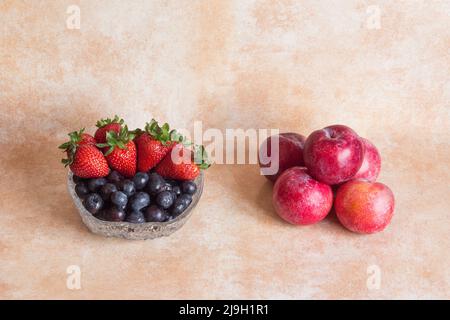 Glasschale mit frischen Erdbeeren und Blaubeeren auf einem abstrakten Hintergrund aus Pastellfarben und einem Haufen roter Pflaumen für hausgemachte Marmelade. Obst und Foo Stockfoto