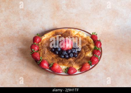 Runder Teller mit köstlichem hausgemachtem Käsekuchen, umgeben von Erdbeeren mit roten Pflaumen und Heidelbeeren in der Mitte auf abstraktem pastellfarbenem Hintergrund Stockfoto