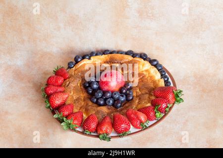 Runder Teller mit köstlichem hausgemachtem Käsekuchen, umgeben von Erdbeeren mit roten Pflaumen und Blaubeeren auf pastellfarbenem Studiohintergrund. Gesund Stockfoto