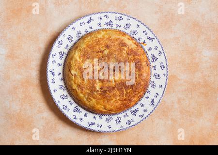 Ansicht von oben traditionelles, hausgemachtes spanisches Kartoffelomelett in einem großen dekorativen Teller mit Blumenmuster auf einem abstrakten pastellfarbenen Studiohintergrund Stockfoto