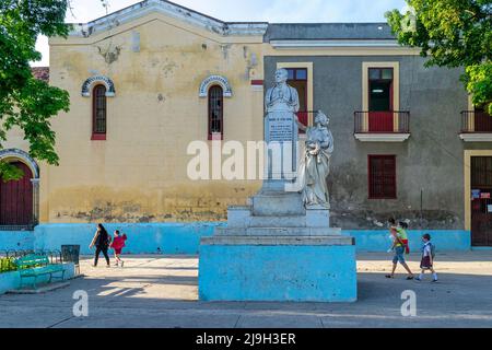 Die Kubaner gehen in einem kleinen öffentlichen Park an einer alten Skulptur von Manuel de Jesus Doval vorbei. Die Parroquia de Jesús, María y José, die eine Kolonialkirche ist Stockfoto