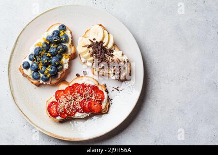 Fruchtige vegetarische Toasts zum Frühstück auf grauem Hintergrund, Draufsicht, Kopierbereich. Brotscheiben mit Ricotta, Beeren, Banane, Schokolade und Samen. Stockfoto