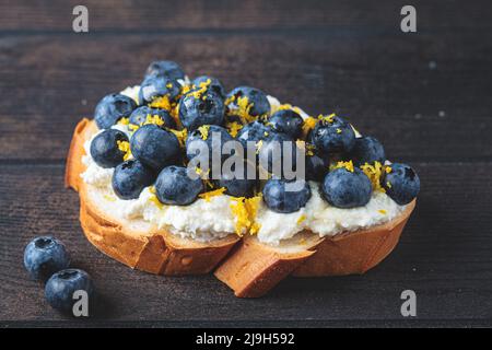 Beerenbrot, vegetarischer Toast zum Frühstück auf dunklem Holzhintergrund, Nahaufnahme. Brotscheibe mit Ricotta, Heidelbeere und Zitrone. Stockfoto