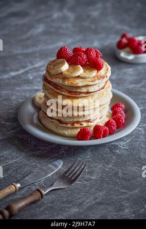 Amerikanische Pfannkuchen mit Himbeeren und Bananen auf dunklem, minimalem Hintergrund. Köstliches Sommerfrühstück auf schwarzem Steintisch. Minimalistisches Design. Stockfoto