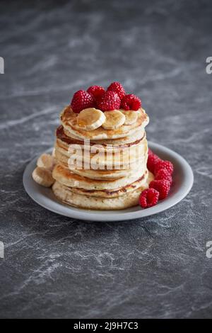 Amerikanische Pfannkuchen mit Himbeeren und Bananen auf dunklem, minimalem Hintergrund. Köstliches Sommerfrühstück auf schwarzem Steintisch. Minimalistisches Design. Stockfoto