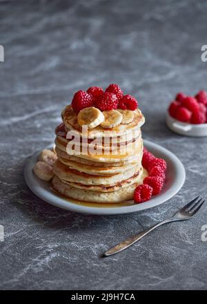 Amerikanische Pfannkuchen mit Himbeeren und Bananen auf dunklem, minimalem Hintergrund. Köstliches Sommerfrühstück auf schwarzem Steintisch. Minimalistisches Design. Stockfoto