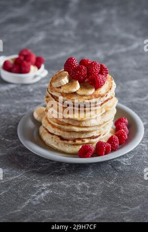 Amerikanische Pfannkuchen mit Himbeeren und Bananen auf dunklem, minimalem Hintergrund. Köstliches Sommerfrühstück auf schwarzem Steintisch. Minimalistisches Design. Stockfoto