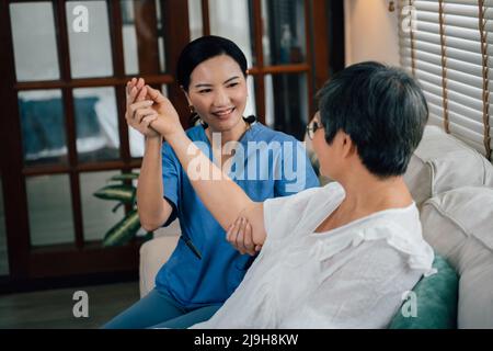 Entzückende weibliche Betreuerin in blauer Uniform lächelnd und streckend Arm einer älteren asiatischen Frau, während sie auf dem Sofa gegen das Fenster im Wohnzimmer zu Hause sitzt Stockfoto