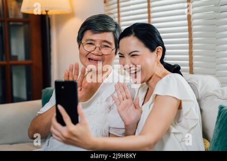 Fröhliche asiatische ältere Mutter und Erwachsene Tochter lächeln und winken die Hände, während sie tagsüber auf dem Sofa sitzen und Videoanruf über Mobiltelefon tätigen Stockfoto